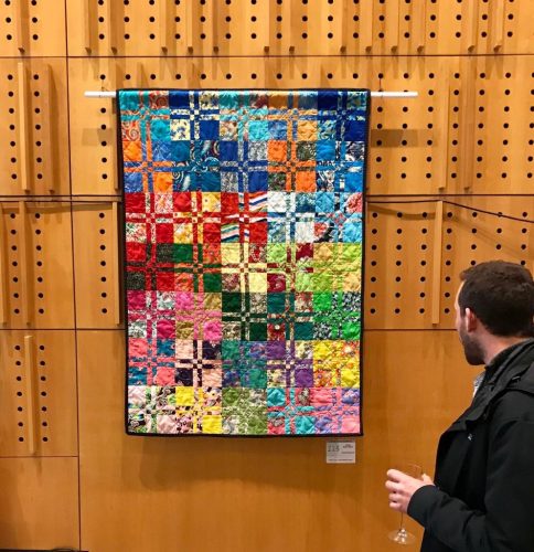 A white bearded man is seen from behind viewing a bright multicolour rectangular quilt hanging on a wood panelled background
