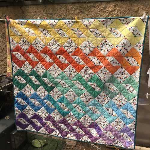 A geometric patterned square quilt, in pastel rainbow colours, hanging on a washing line