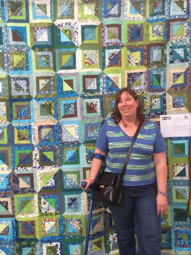 Alison is standing in front of a large blue and green quilt, using a crutch